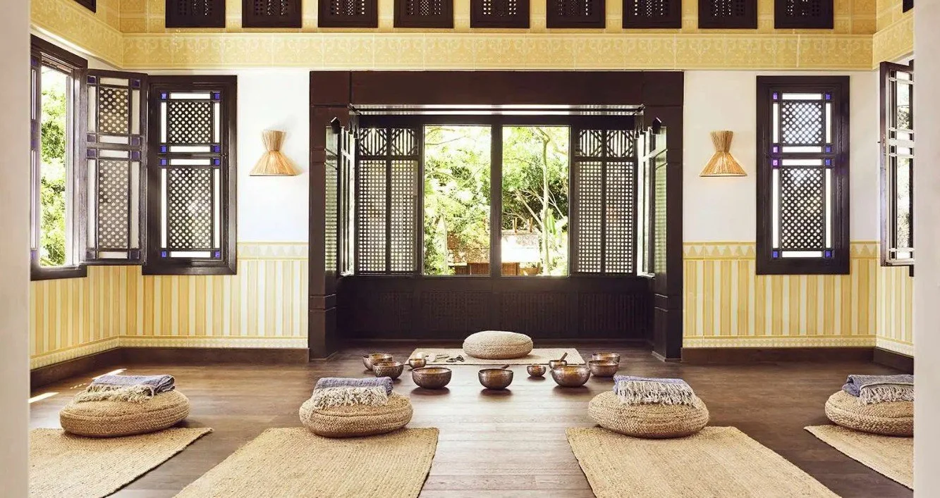 A yoga or meditation room with open windows, wooden floors, and banquettes. Cushions and bowls are arranged on the floor for meditation or relaxation.