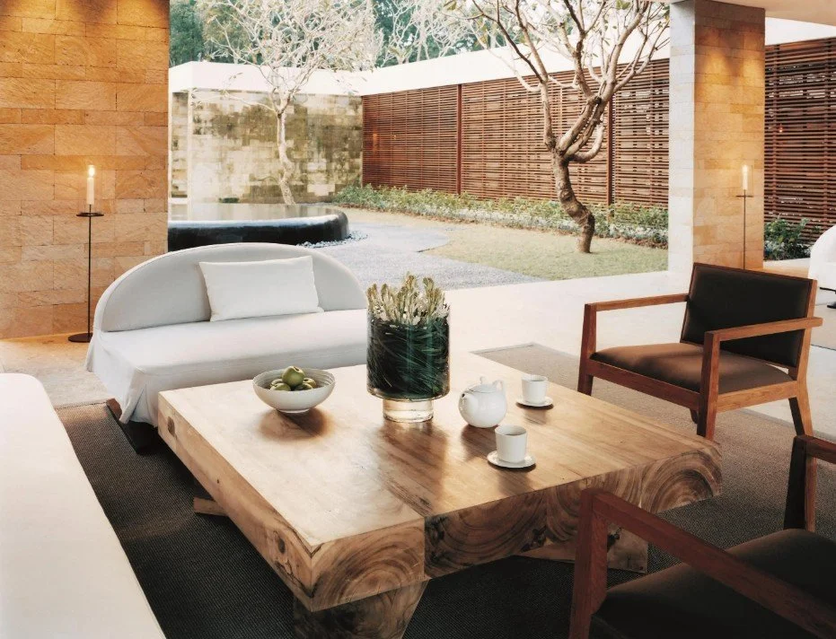 Modern living room with white and brown furniture, a large wooden coffee table, and an open view of a backyard with trees and a stone water feature.