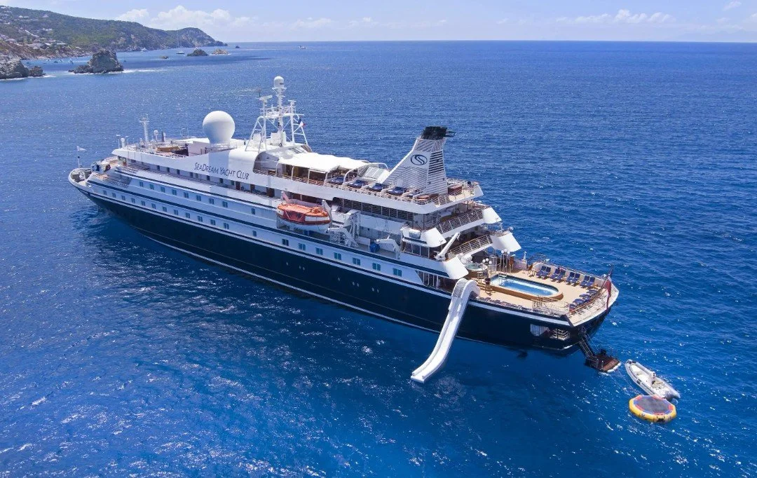 A large white yacht with a slide and swimming pool on deck sailing in blue ocean waters near a rocky coastline.