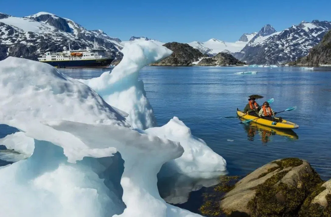 Kayakers paddling near icebergs in a cold, mountainous, icy landscape with a large ship in the background.