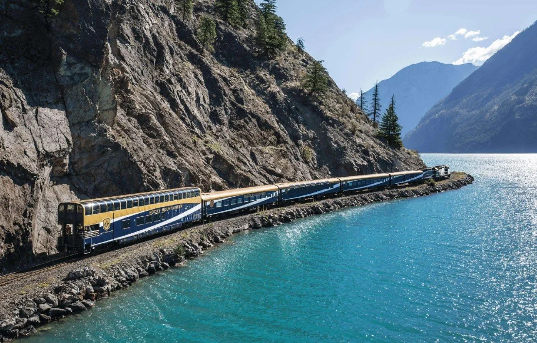 A passenger train labeled 'Rocky Mountaineer' traveling along a narrow rocky pathway beside a large body of water, with mountains in the background.