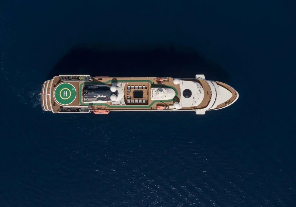 Top-down aerial view of a yacht sailing in dark blue water, showing the helipad, deck, and bow.