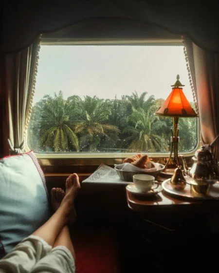 Person relaxing by a window with a view of palm trees, sitting on a bed or chair with feet up, surrounded by a lamp, teapot, cups, and snacks.