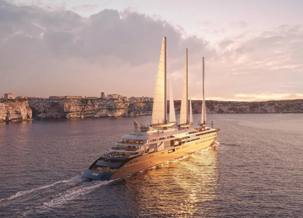 Luxury sailboat with multiple masts sailing near rocky cliffs at sunset.