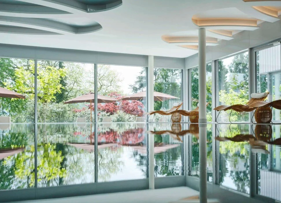 Modern indoor swimming pool with large glass windows showing lush greenery outside, wicker lounge chairs, and patio umbrellas.