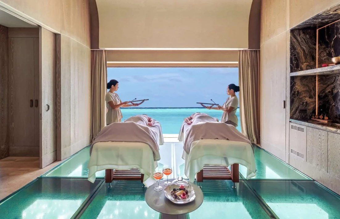 Two women receiving massages in a spa room with a large window overlooking the ocean, with a table of drinks and snacks in the foreground.