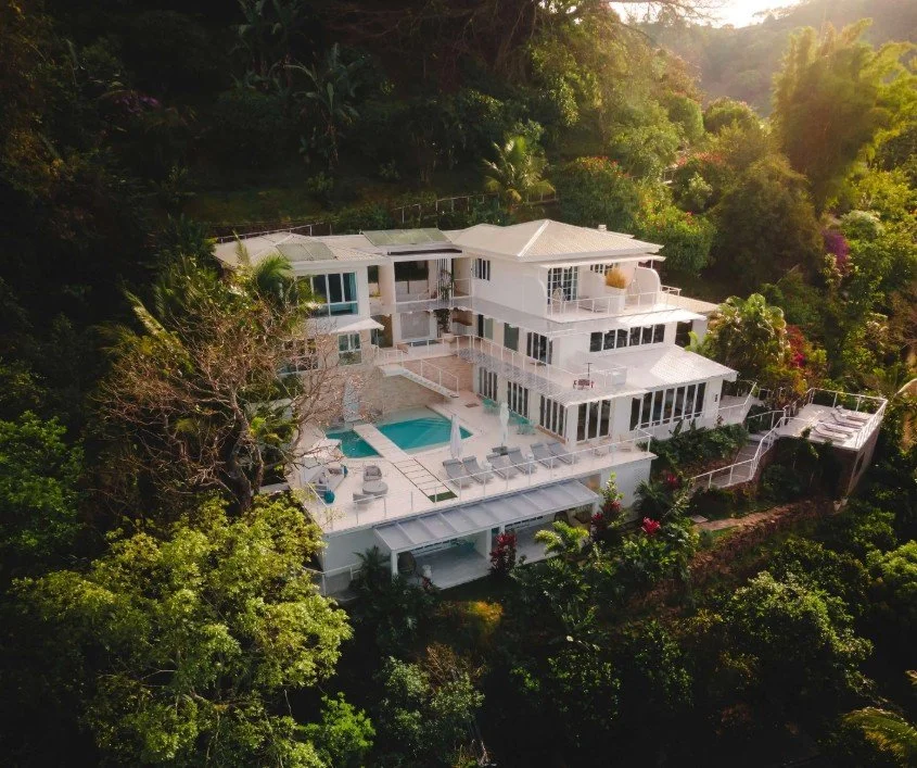A large white multi-story house with multiple balconies, a pool, and outdoor seating, situated on a hillside surrounded by lush green trees.