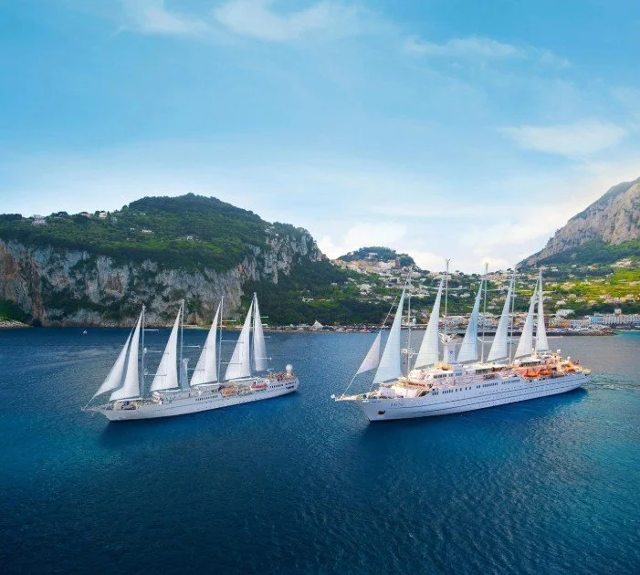 Two large white sailboats with multiple sails sailing near a mountainous coastline under a partly cloudy sky.