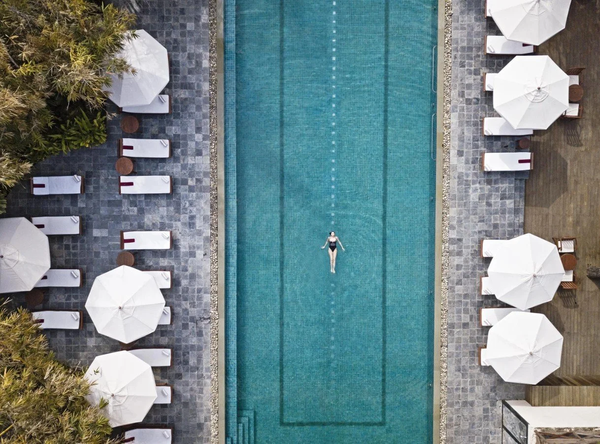 An aerial view of a swimming pool with a person floating on their back in the center. The pool is surrounded by lounge chairs with white umbrellas and shaded sitting areas on a stone patio.