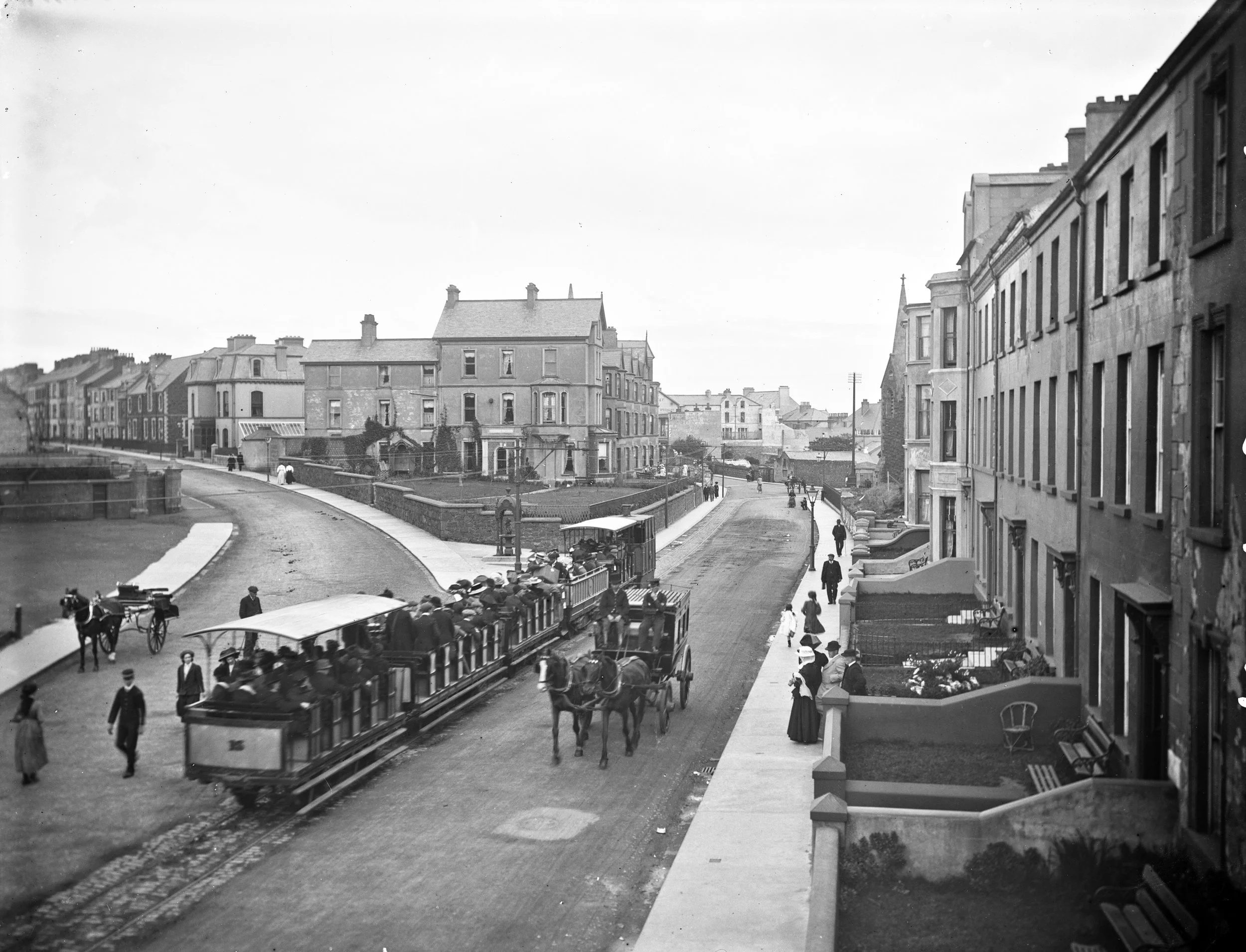A black-and-white photo of a vintage street scene with a streetcar carrying passengers, horse-drawn carriages, and pedestrians walking on sidewalks.