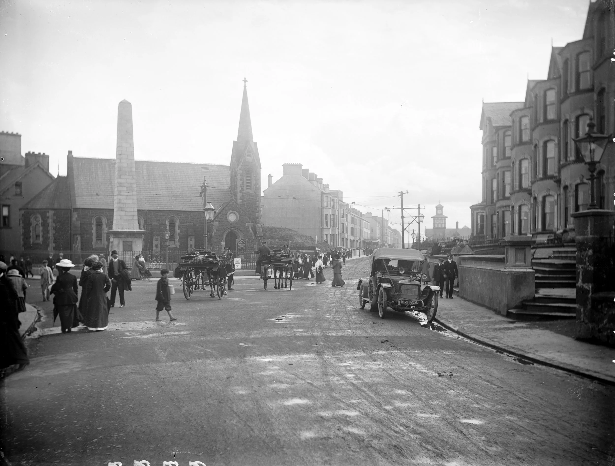 A historical black and white photo of a busy early 20th-century street scene with pedestrians, horse-drawn carriages, a vintage automobile, and buildings including a church with a tall steeple.