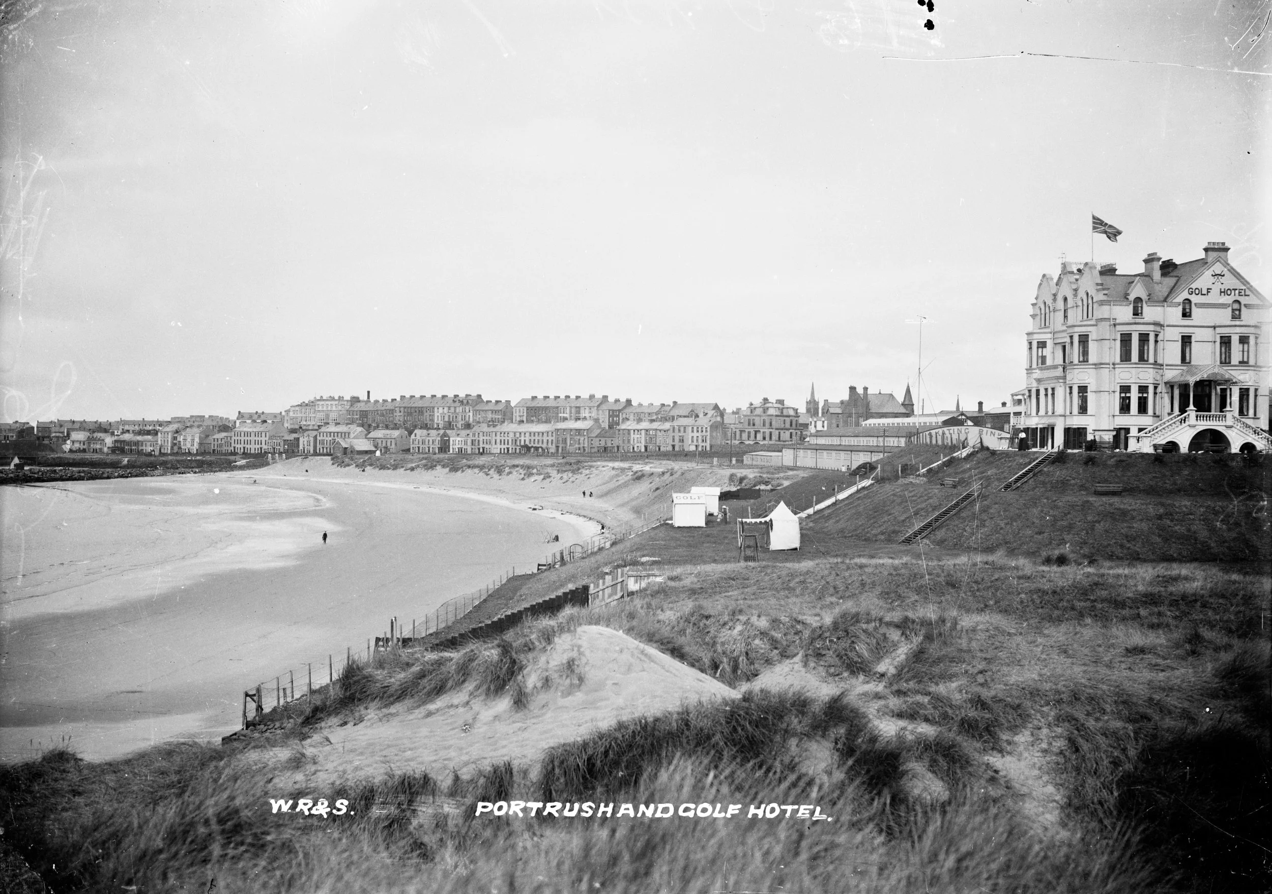 Historical black and white photograph of the Portrush and Golf Hotel, showing a beach and hotel building with the sea in the foreground and town buildings in the background.