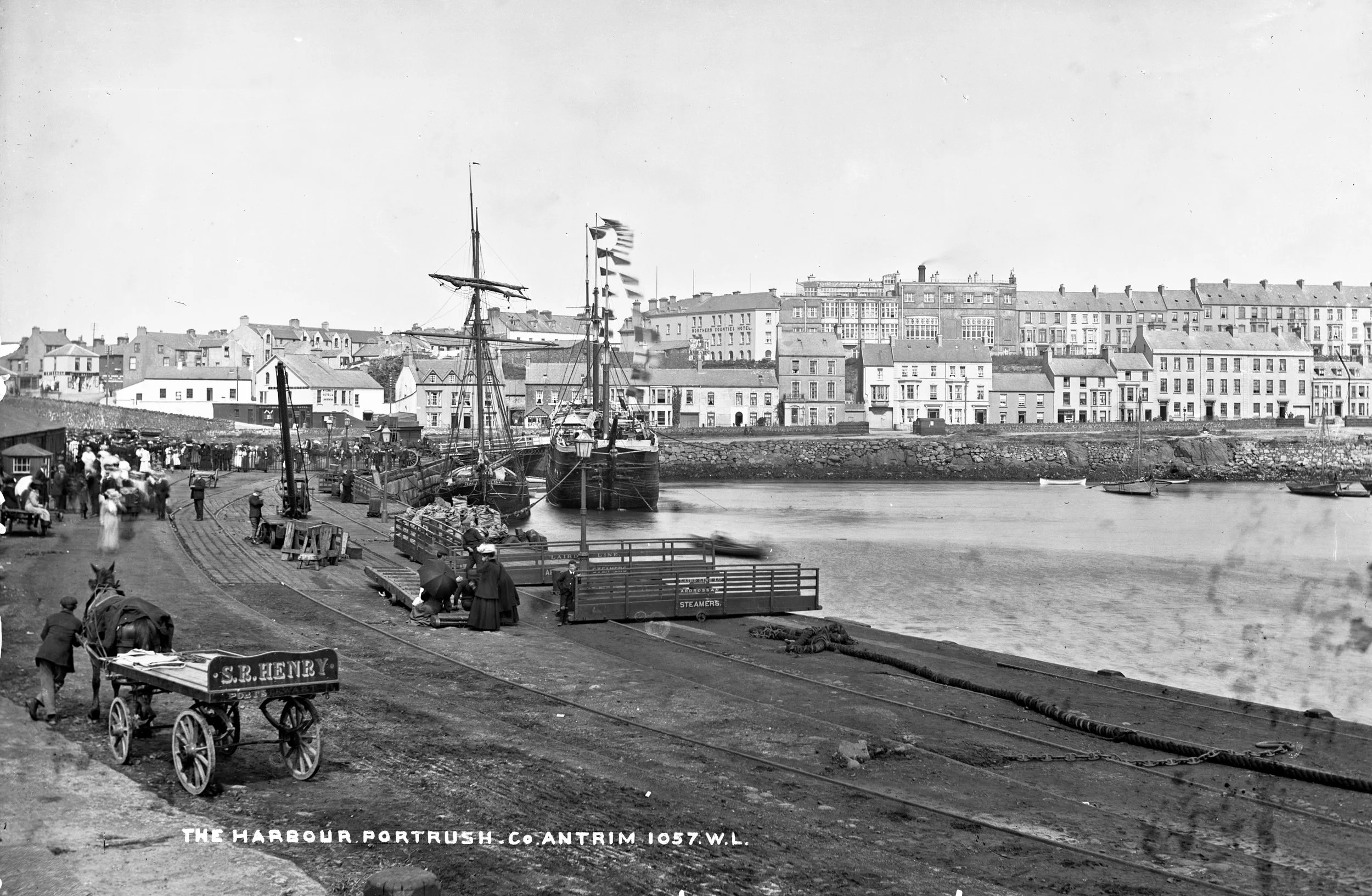 Black and white photo of a historic harbor with ships, buildings, and people along the waterfront.