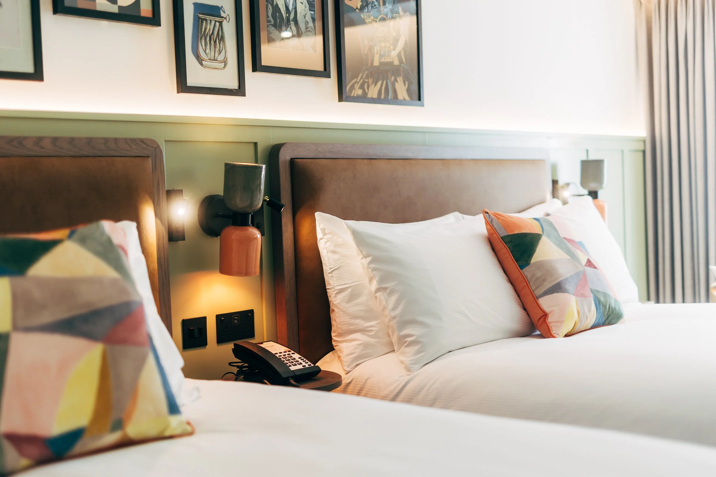 Two neatly made beds with colorful geometric pillows in a hotel room, featuring wall-mounted lamps and framed artwork above the headboards.