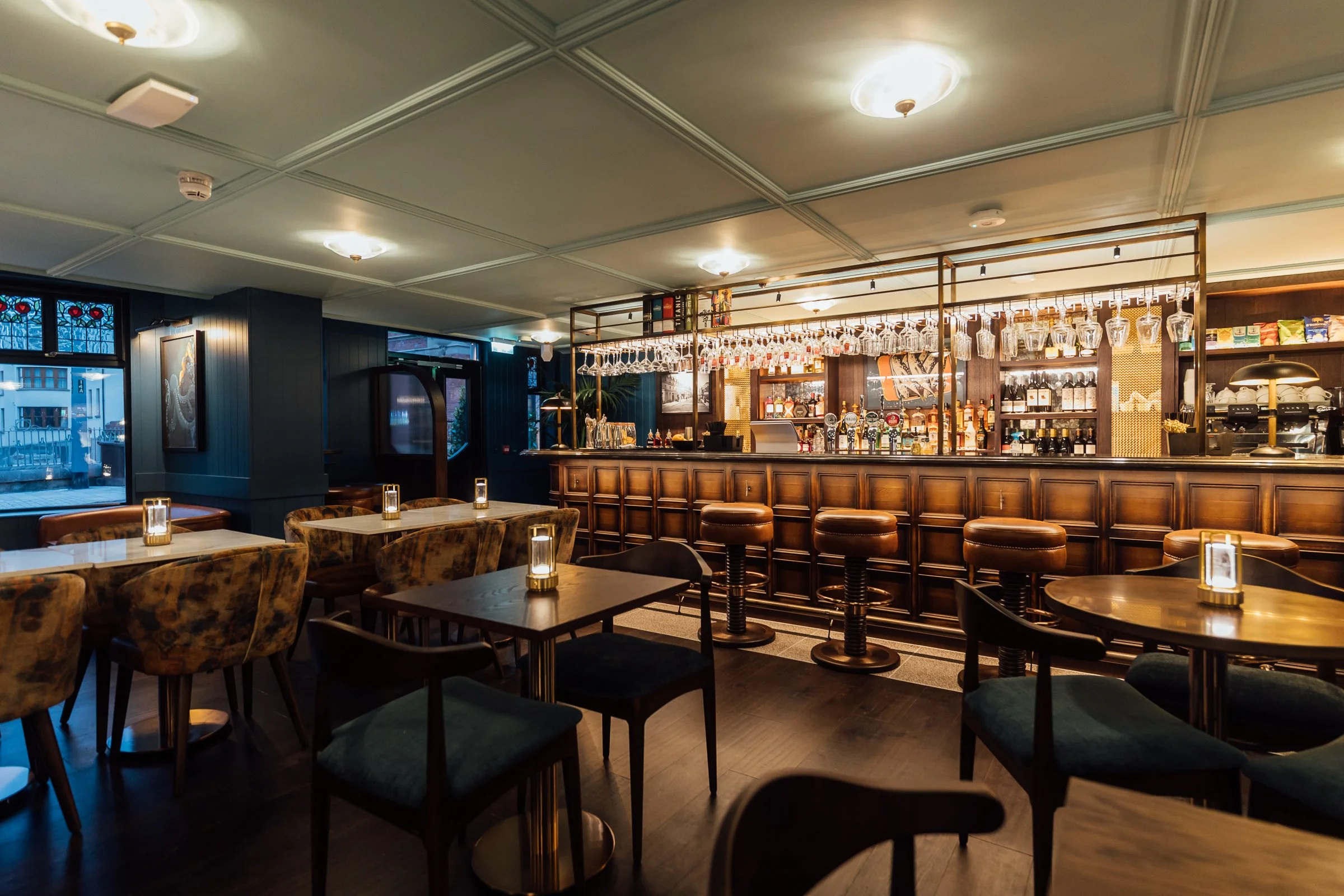 A cozy bar and restaurant interior with wooden bar counter, bartender area, shelving with liquor bottles, hanging wine glasses, dark chairs, illuminated candlelit tables, and windows with stained glass accents.