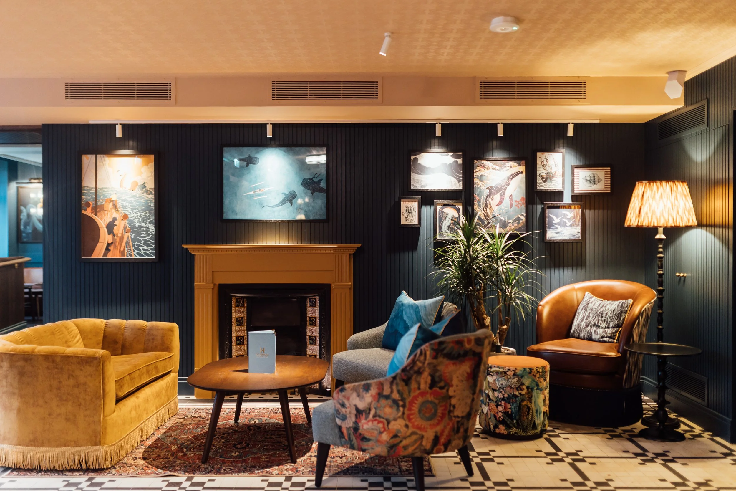A cozy living room with dark panel walls decorated with framed artwork, including underwater scenes. There is a yellow velvet sofa, a patterned armchair, a gray loveseat with pillows, a round ottoman with floral upholstery, a potted plant, a wooden c
