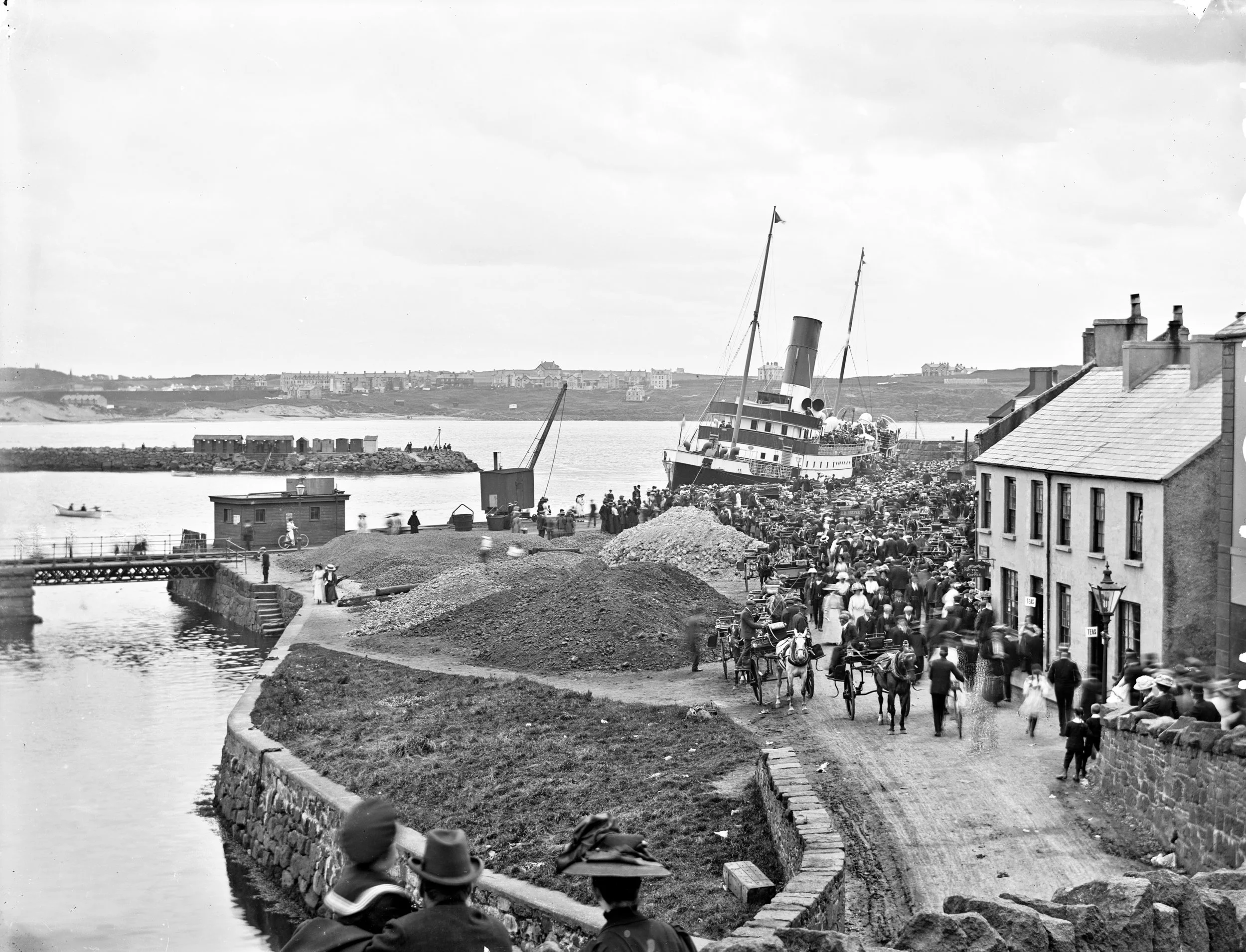 A crowded dock along the waterfront with a large ship tilted on the shoreline and numerous people walking, riding bicycles, and arriving in horse-drawn carriages, with buildings lining the street and a body of water in the background.