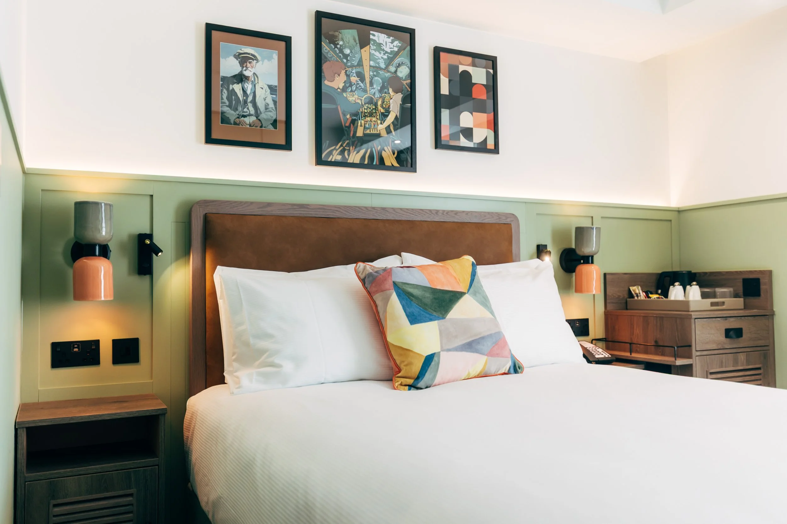 A neatly made hotel bed with white bedding, a colorful geometric pillow, and a brown headboard. The wall behind features three framed artworks, and there are modern wall-mounted lamps on each side of the bed. A wooden nightstand is on the left, and a