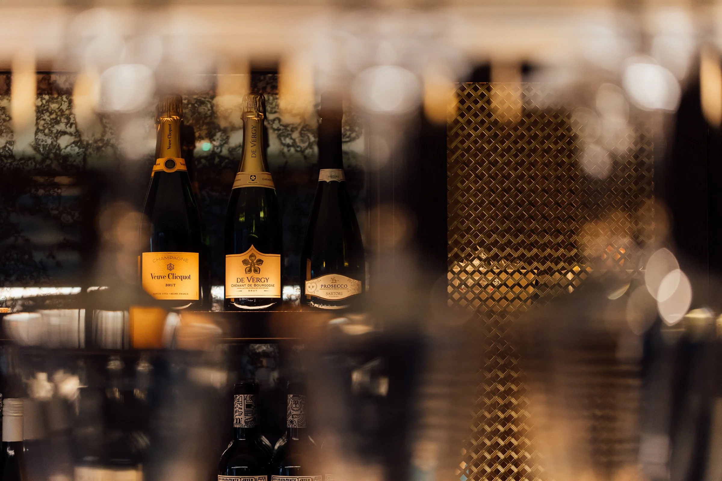 Three bottles of champagne and sparkling wine on a wine shelf, viewed through a blurred foreground of other bottles, with warm lighting.