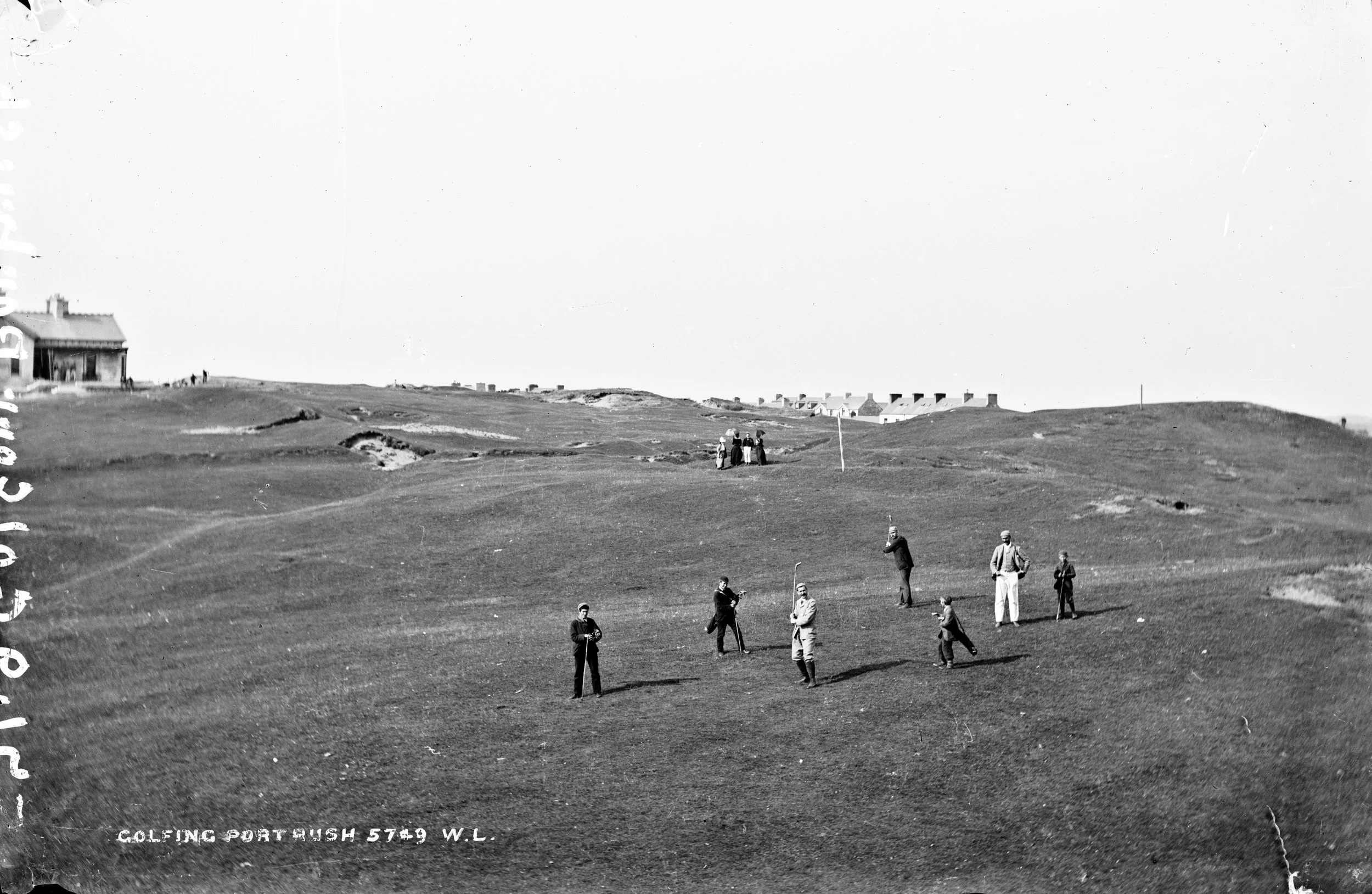Group of people playing golf on a grassy golf course with houses in the background, black and white photo from early 20th century.
