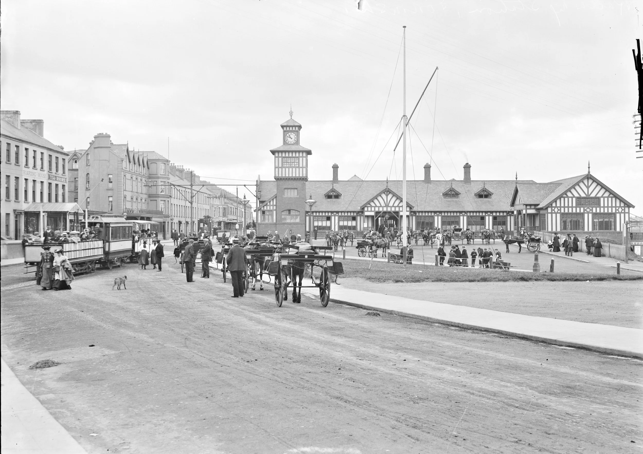 Historical black and white photo of a busy train station platform with people, horses, carriages, and a clock tower in the background.
