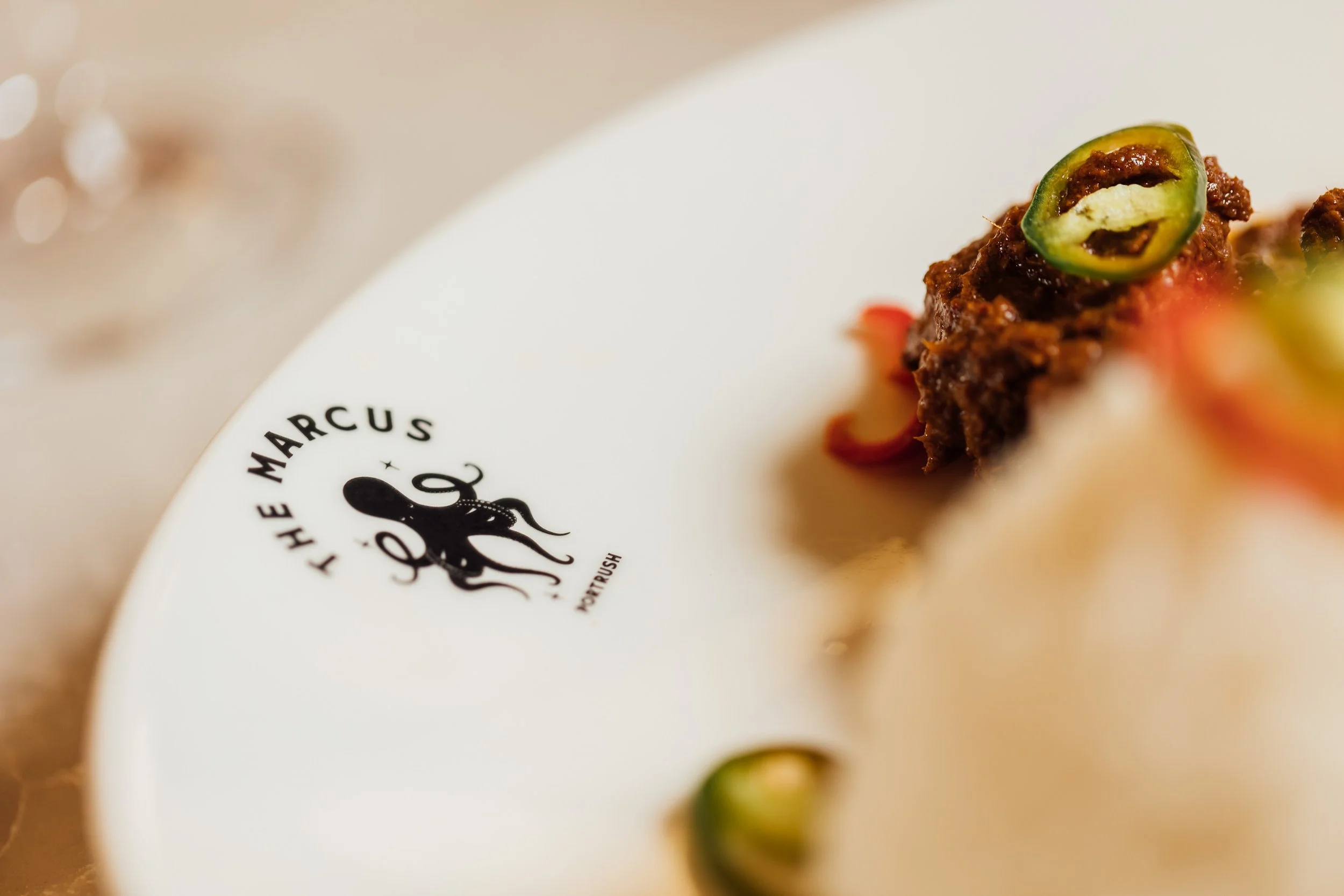 A close-up of a white plate with the black logo 'The Marcus' and an octopus illustration, featuring a piece of food topped with green chili slices and garnished with red sauce.