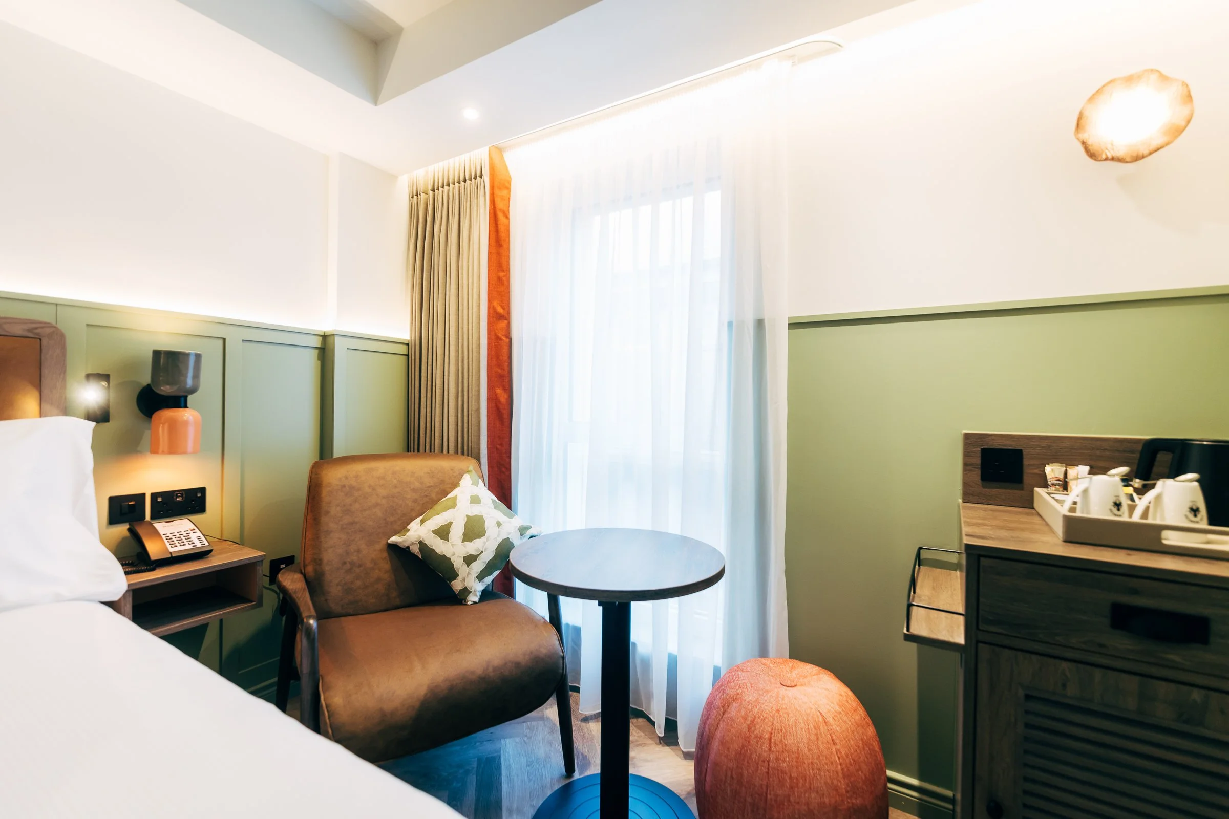 Hotel room with a bed, a small round table, an armchair with a pillow, a window with curtains, and a cabinet with tea and coffee set.