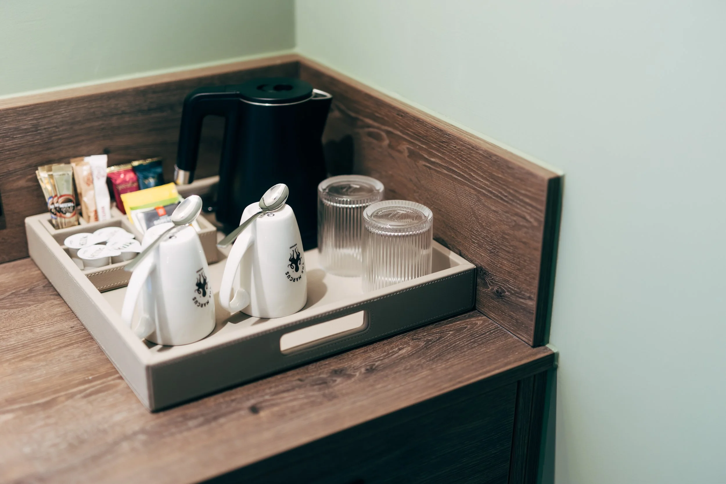Tray with coffee supplies including coffee packet, creamer, sugar, glass cups, and electric kettle on wooden surface