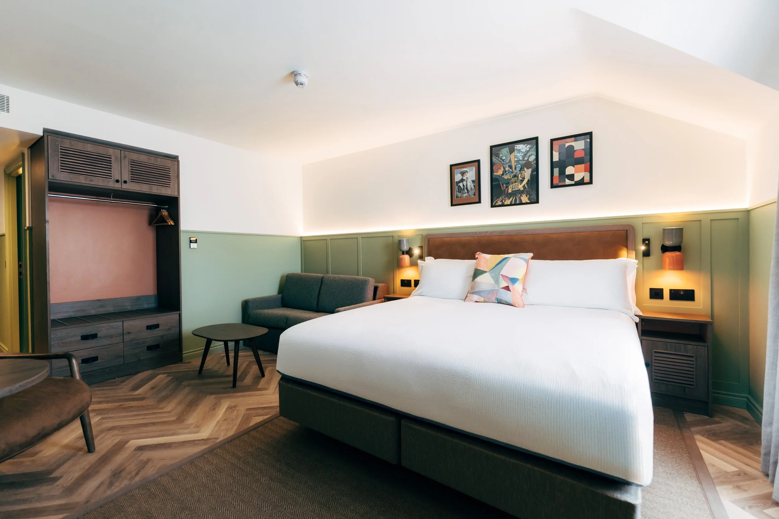 Modern hotel room with a large bed, colorful pillows, green and white walls, framed artwork, bedside lamps, a sofa, a small round table, and a wooden open wardrobe.