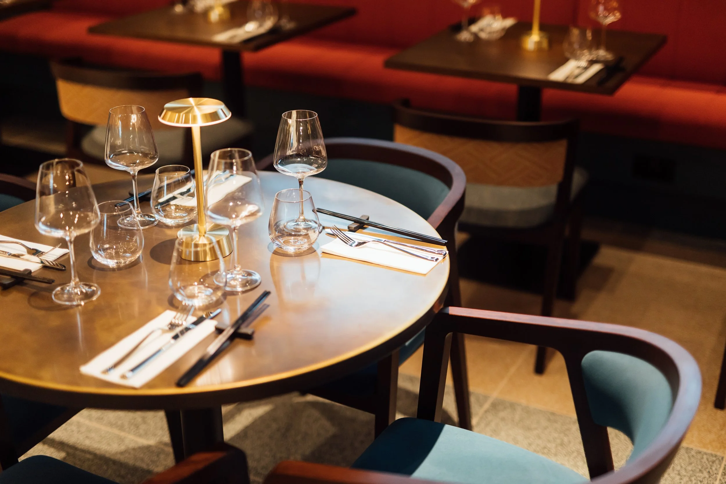 A round wooden dining table set for four with wine glasses, chopsticks, forks, knives, and napkins, illuminated by a small gold table lamp, in a dimly lit restaurant with empty chairs and booths in the background.