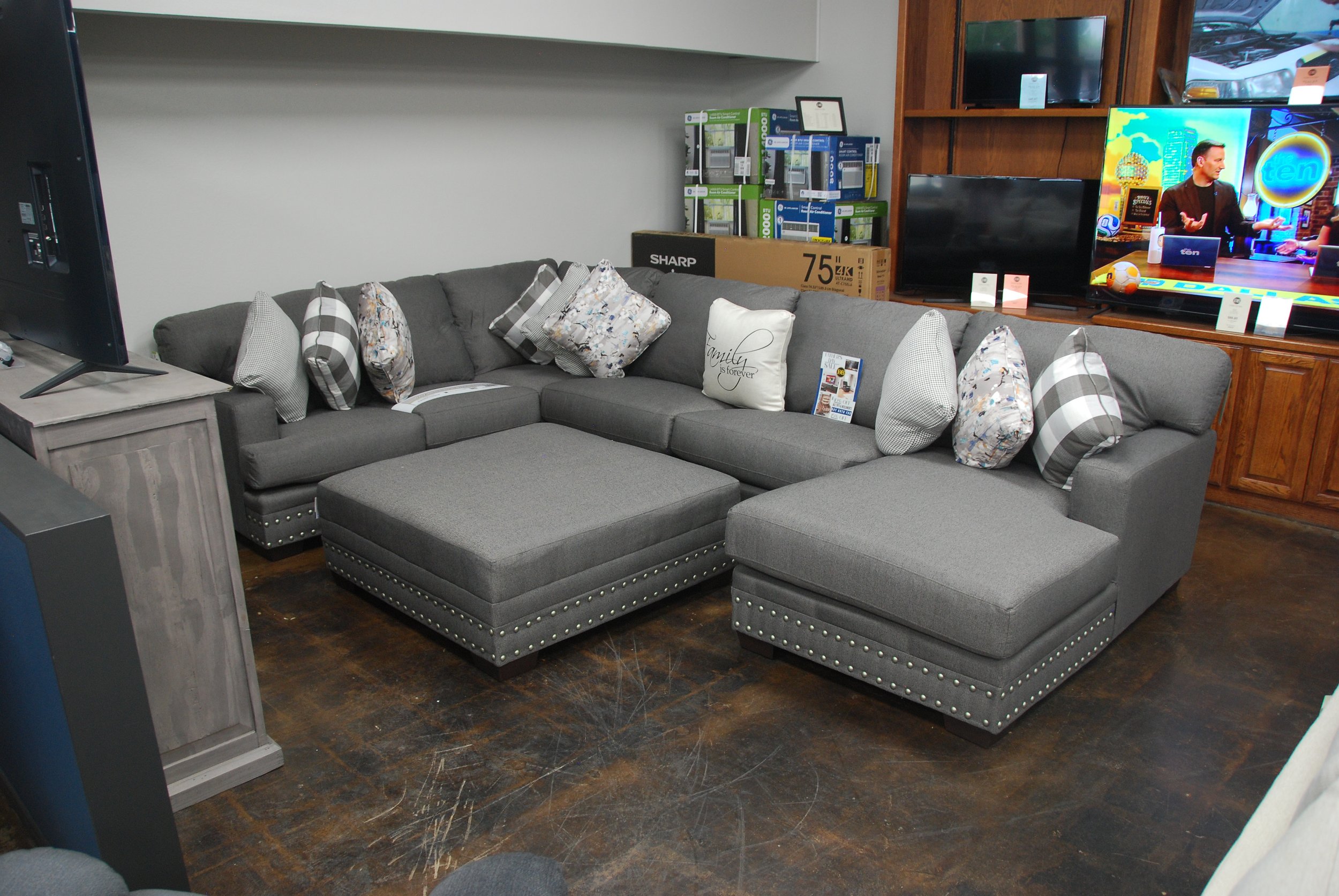 A light gray sectional sofa with several decorative pillows in neutral tones, situated on a dark wood floor in a furniture store.