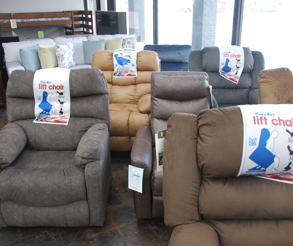 Two recliner chairs, one beige and one dark gray, displayed in a furniture store with signs on top indicating they are lift chairs.