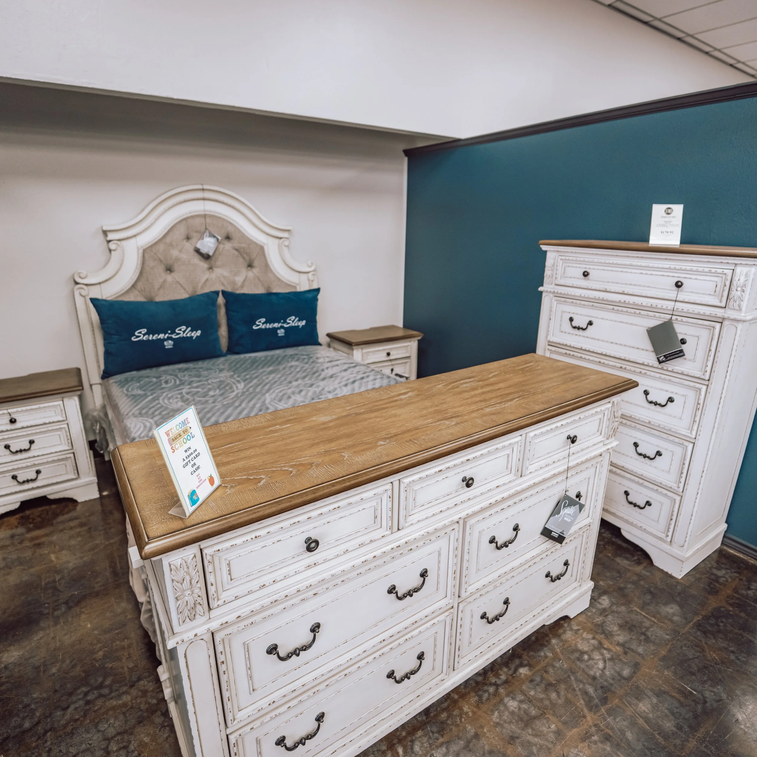 A bedroom furniture set displayed in a furniture store, including a white bed with a decorative headboard, two white nightstands, and a white dresser with multiple drawers. There are blue pillows on the bed with white text, and the dresser has price tags hanging from the handles. The room has a blue accent wall and a wood-look floor.