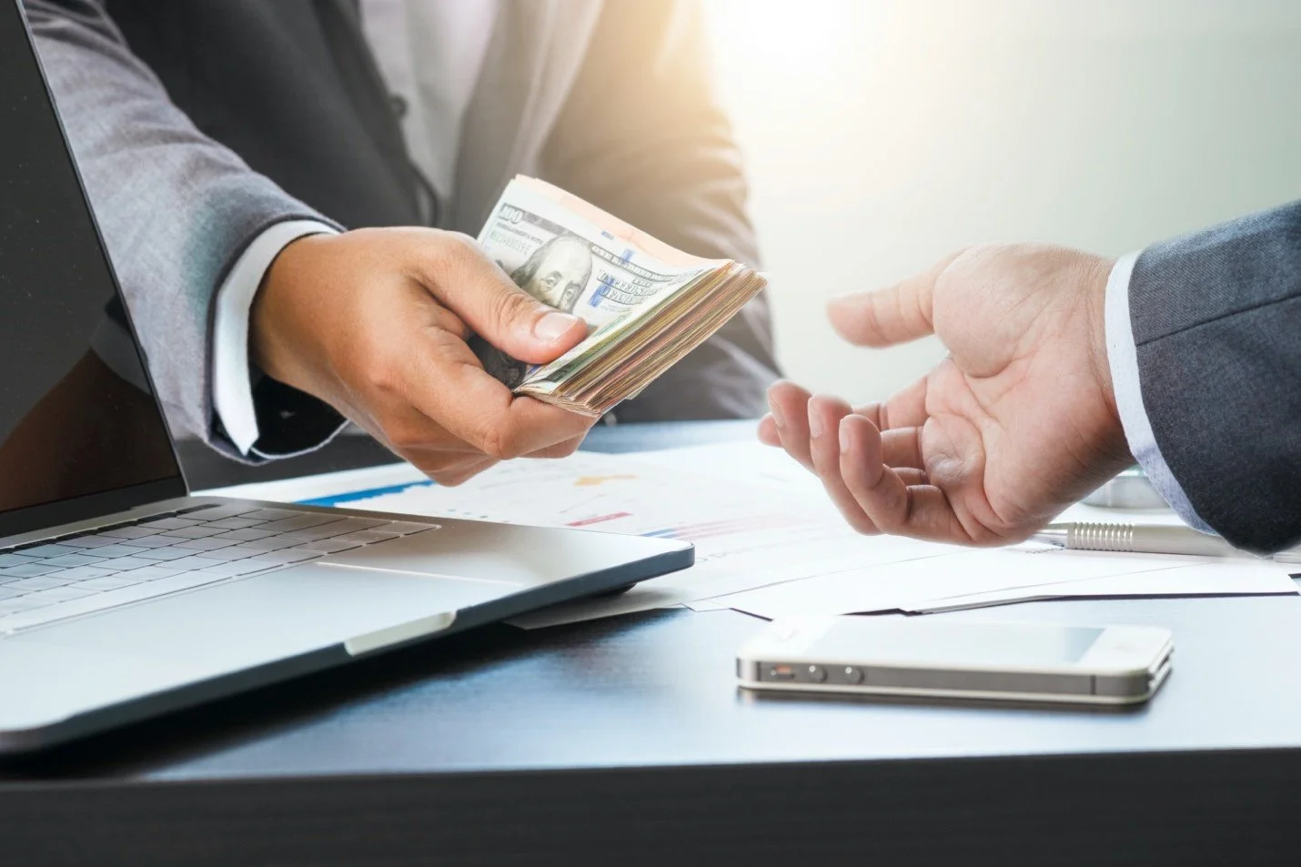 Business person handing over a bundle of US dollar bills to another person at a desk with a laptop, smartphone, and papers.