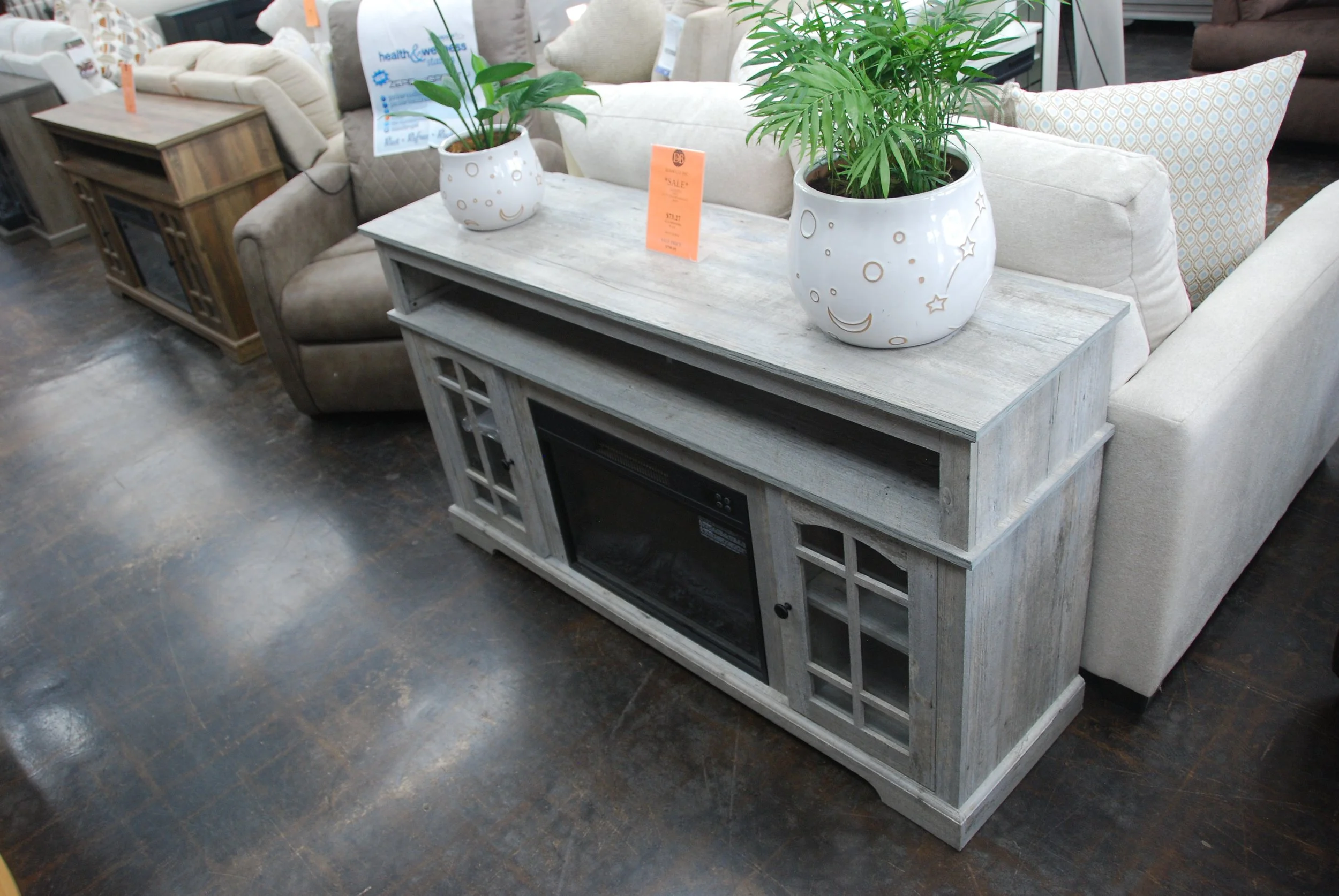 A gray wooden TV stand with glass-paneled cabinet doors, a built-in electric fireplace, and open shelving, situated in a furniture store among various sofas and chairs.