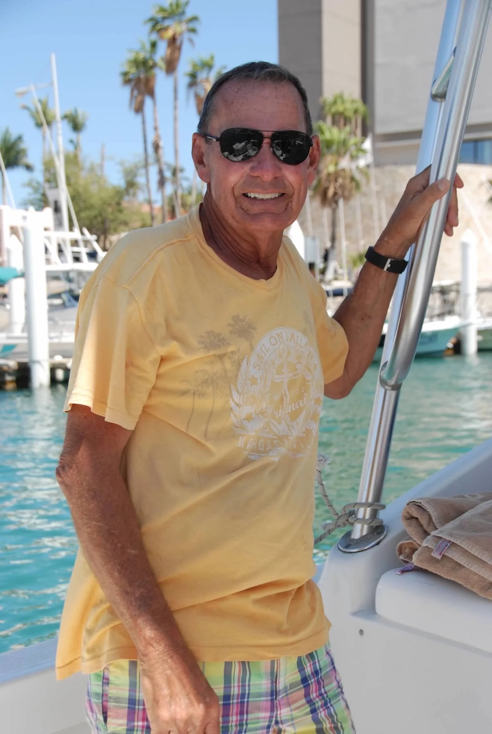Older man wearing sunglasses, a yellow t-shirt, and plaid shorts, smiling and holding onto a boat ladder at a marina with palm trees and boats in the background.