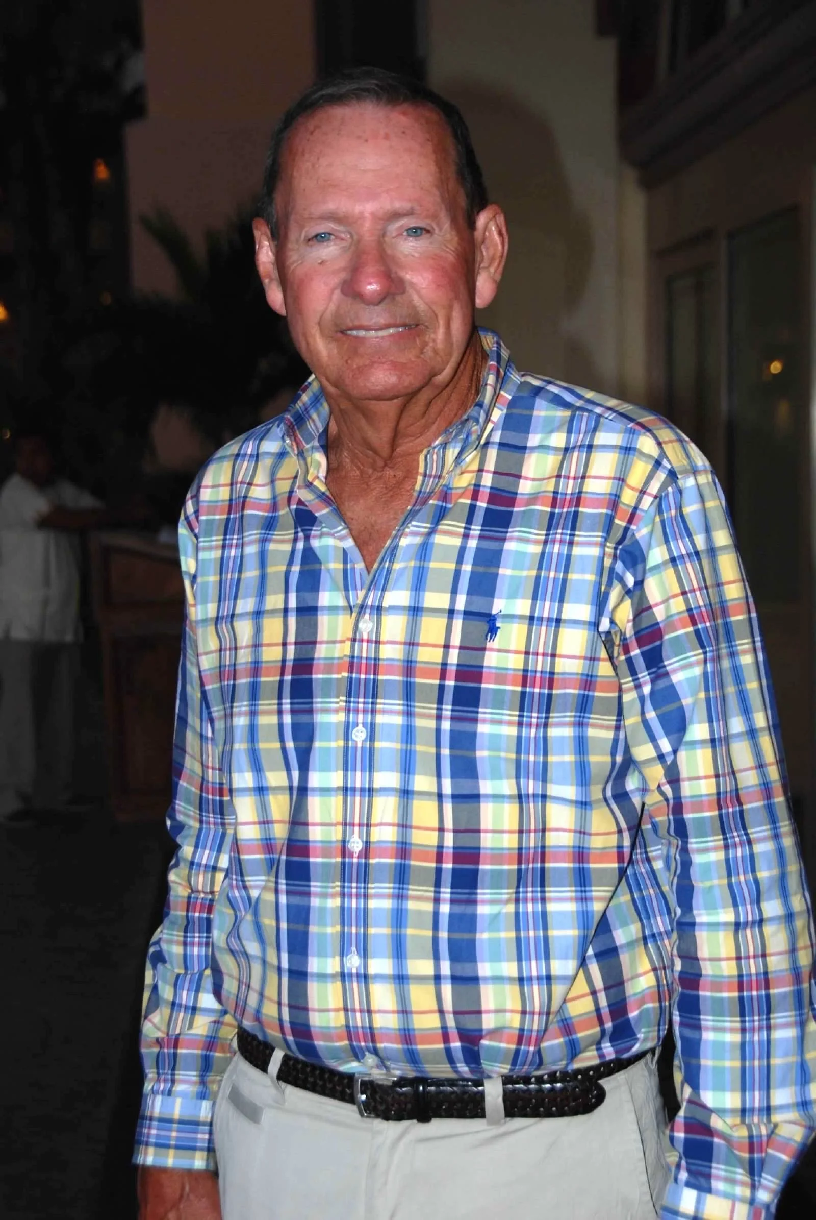 A middle-aged man with blue eyes and short dark hair, wearing a colorful plaid button-up shirt and light-colored pants, standing indoors during evening time.