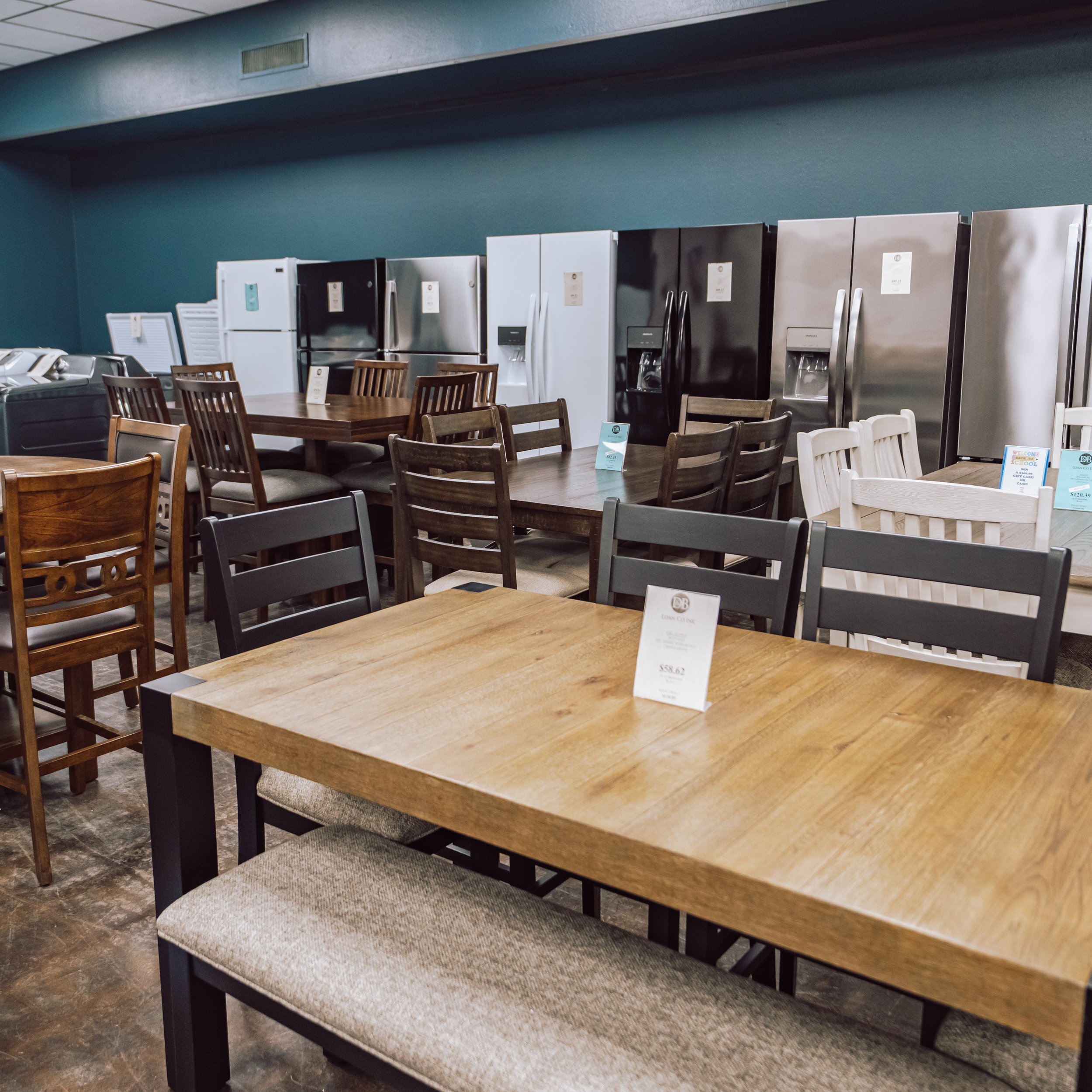 Display of various refrigerators and kitchen appliances in a furniture store with tables and chairs in the foreground.