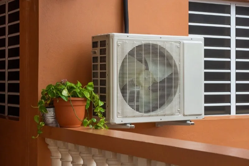 Air conditioning unit mounted on a balcony with a potted plant nearby, against an orange wall and metal window shutters.