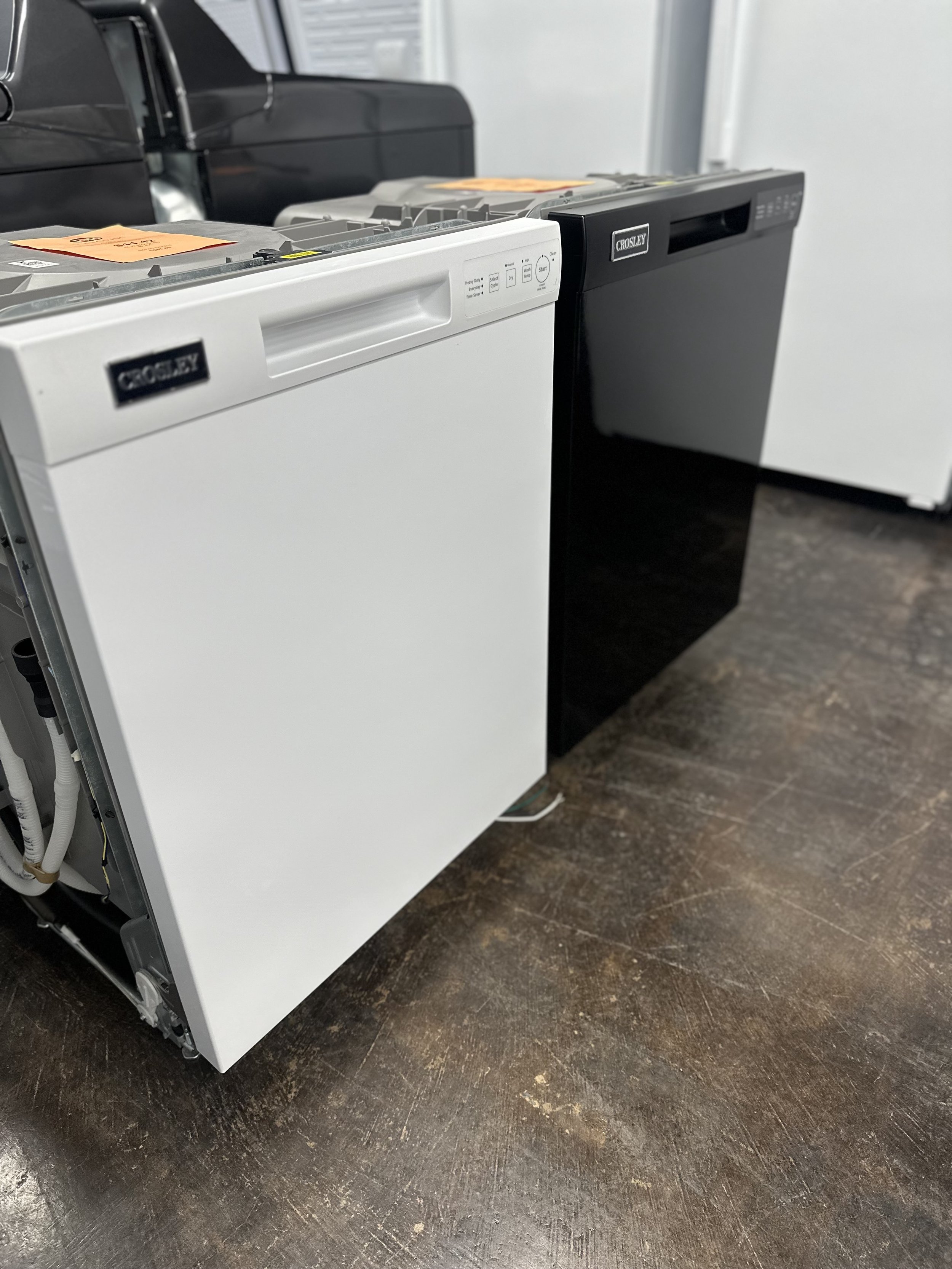 Two dishwashers, one white and one black, placed side by side on a dark wooden floor.