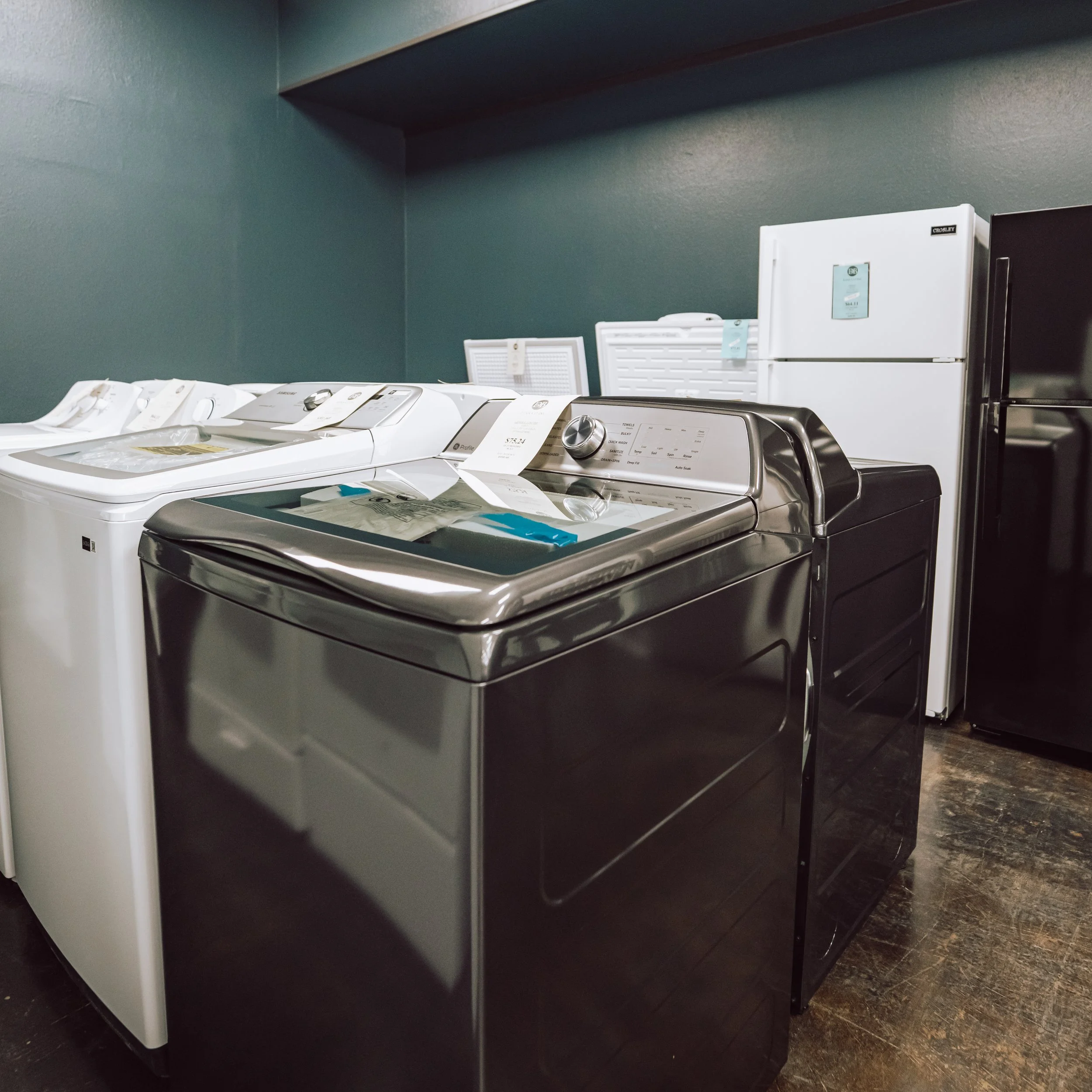 A showroom with two washing machines and two refrigerators on a wooden floor against a dark green wall.
