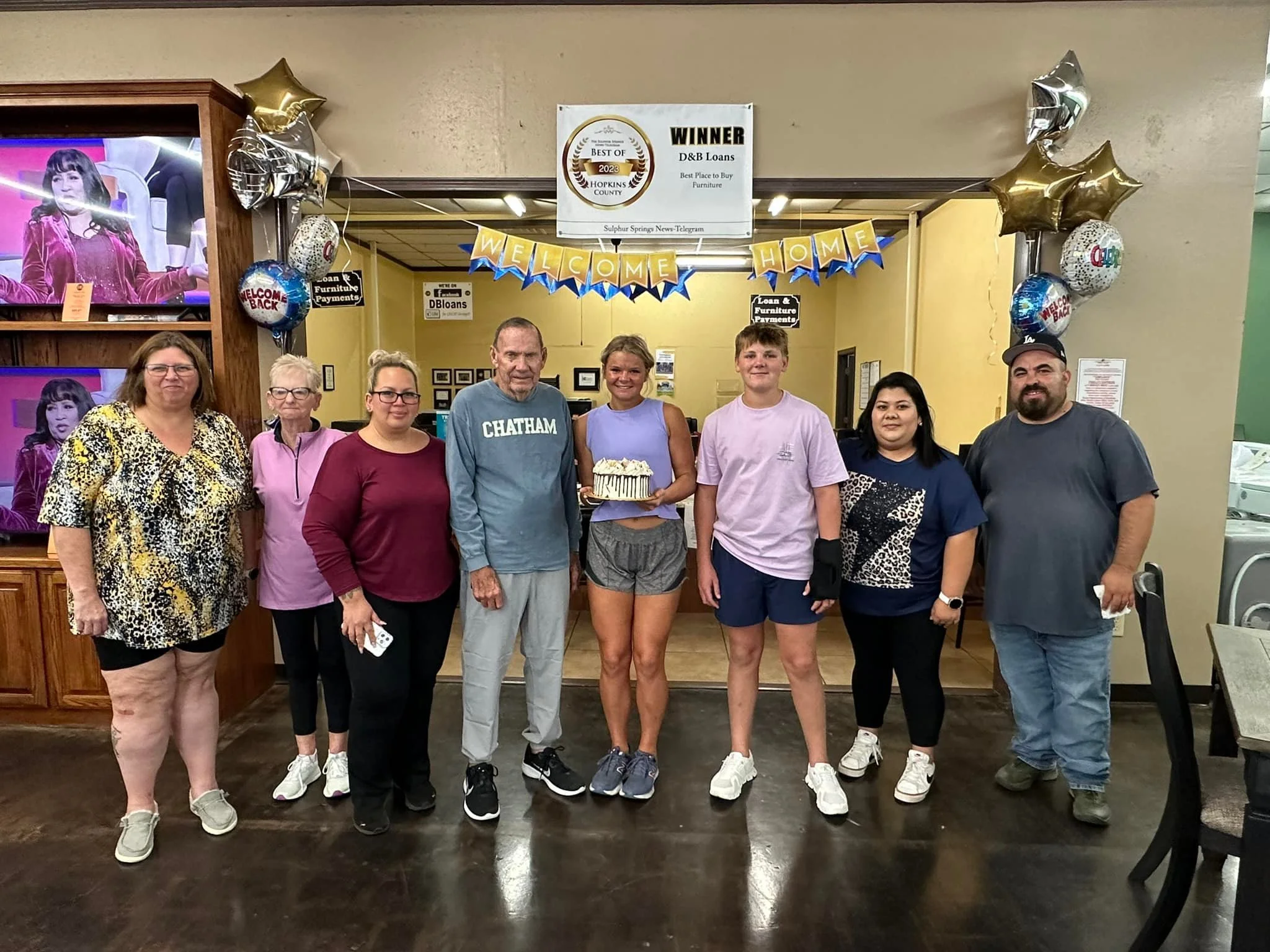 Group of nine people standing in a restaurant or cafe, with a woman holding a cake in the center, celebrating a special occasion, with balloons and a 'Welcome Home' banner in the background.