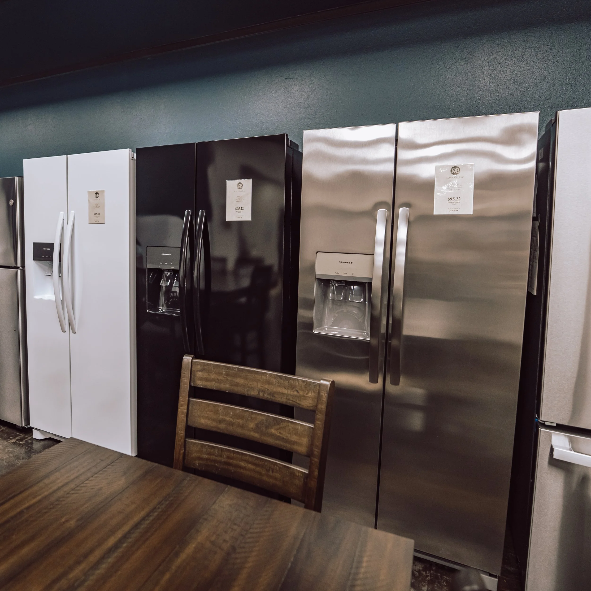 A row of refrigerators in various colors and finishes, including white, black, and stainless steel, displayed in a store showroom.