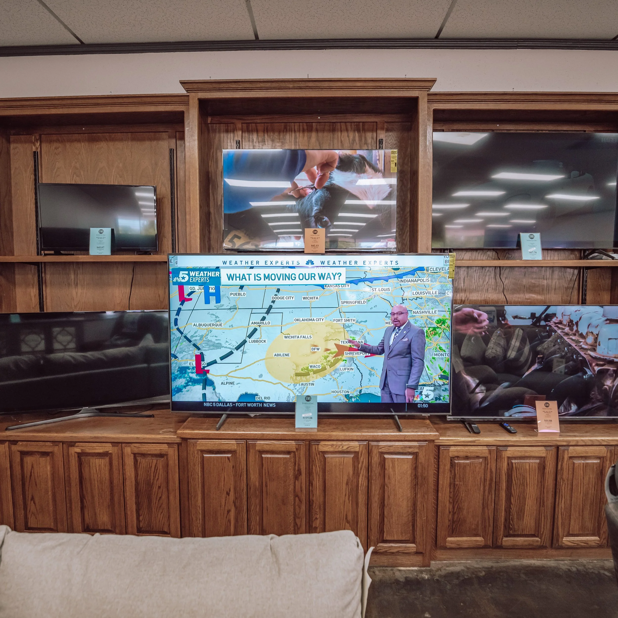 A store display with multiple flat-screen TVs, with a prominent large screen showing a weather forecast segment. The weather forecast features a weather presenter pointing at a map, with a headline reading 'WHAT IS MOVING OUR WAY?'. Other screens are turned off or displaying different images, and there's a wooden shelf and cabinetry beneath the TVs.
