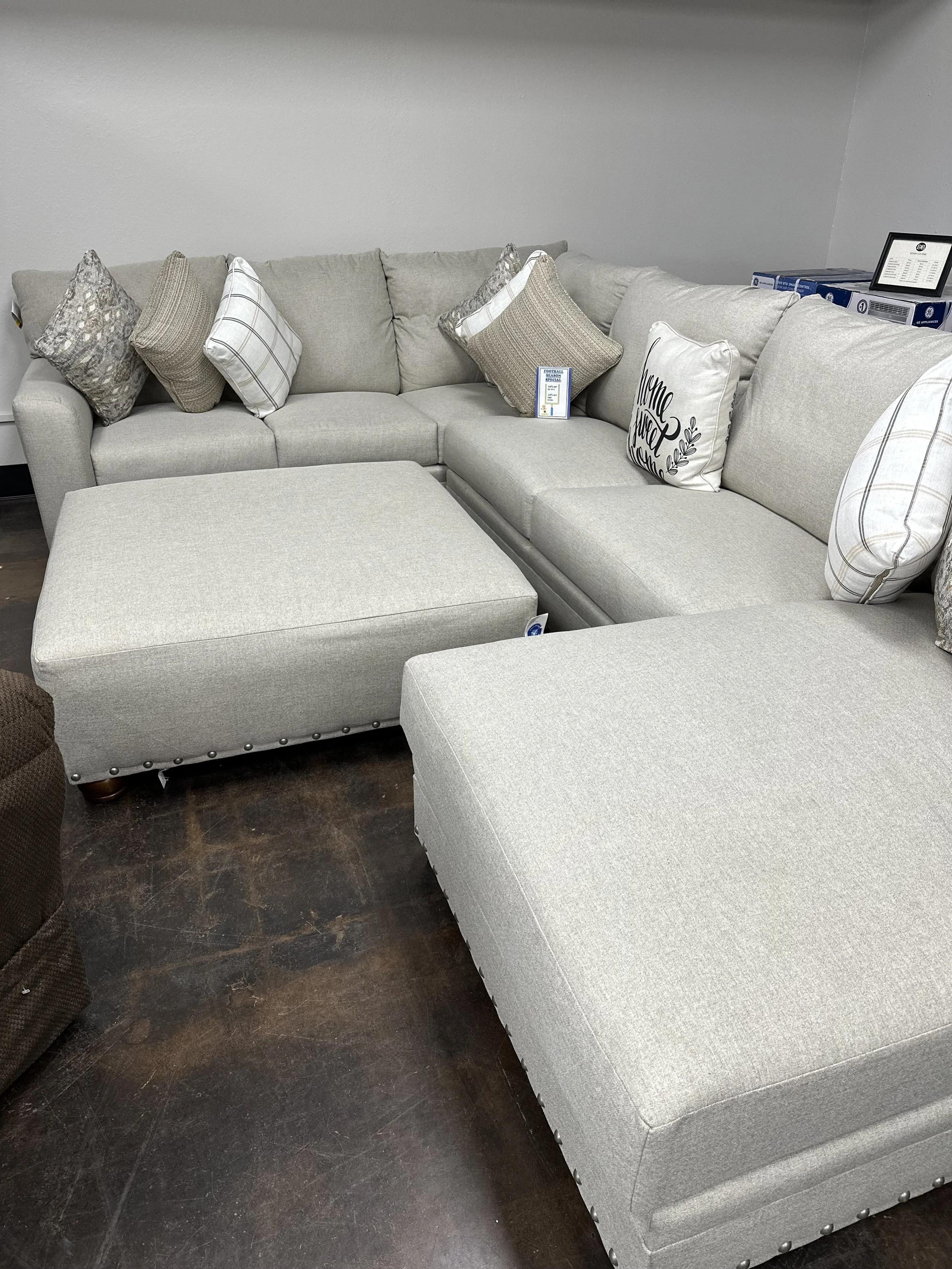 A light gray sectional sofa with several decorative pillows in neutral tones, situated on a dark wood floor in a furniture store.