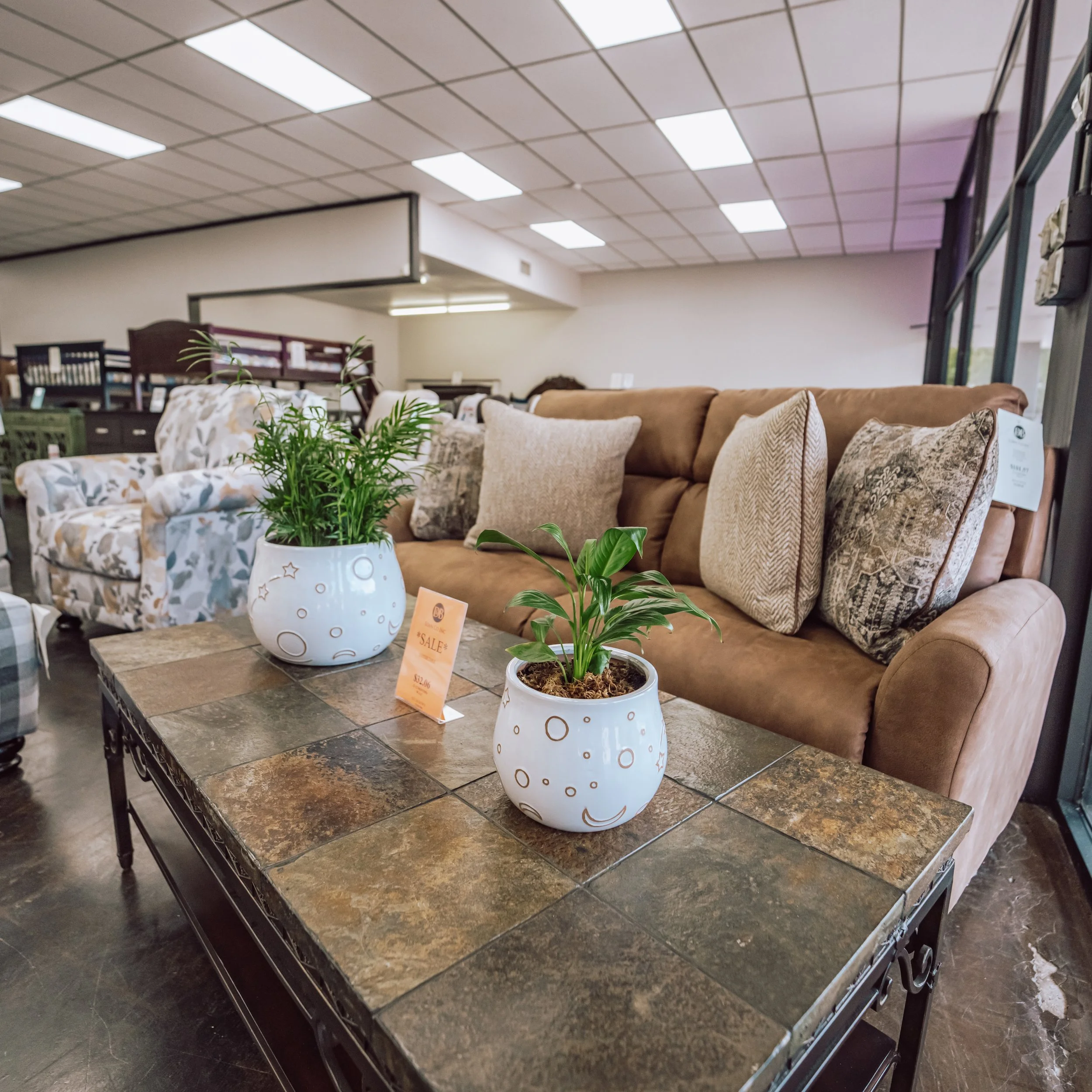 Showroom of living room furniture with a brown sofa, decorative pillows, and potted plants on a tiled coffee table.