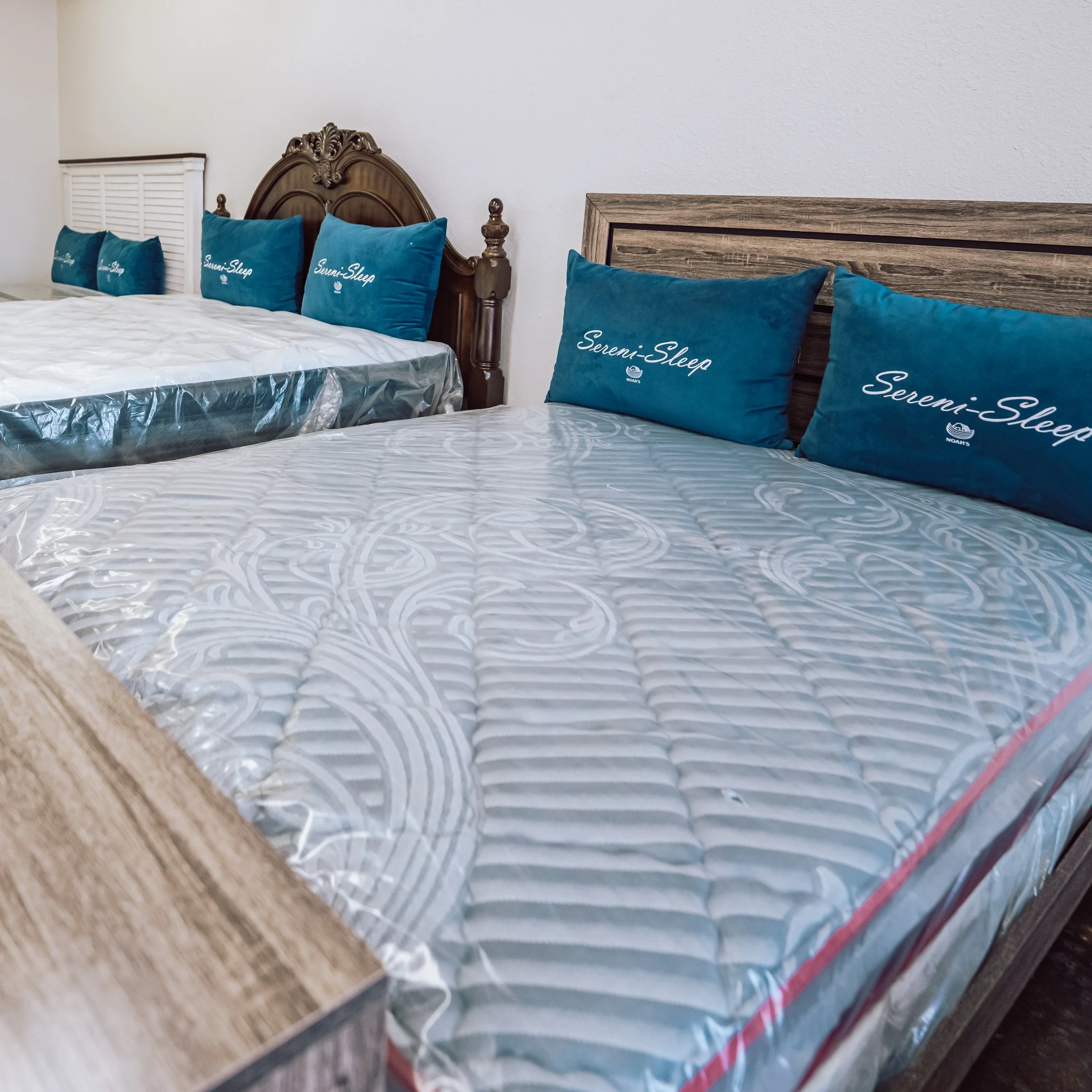 Two beds with mattresses and blue pillows that have 'Sereni-Sleep' written on them, in a room with white walls.