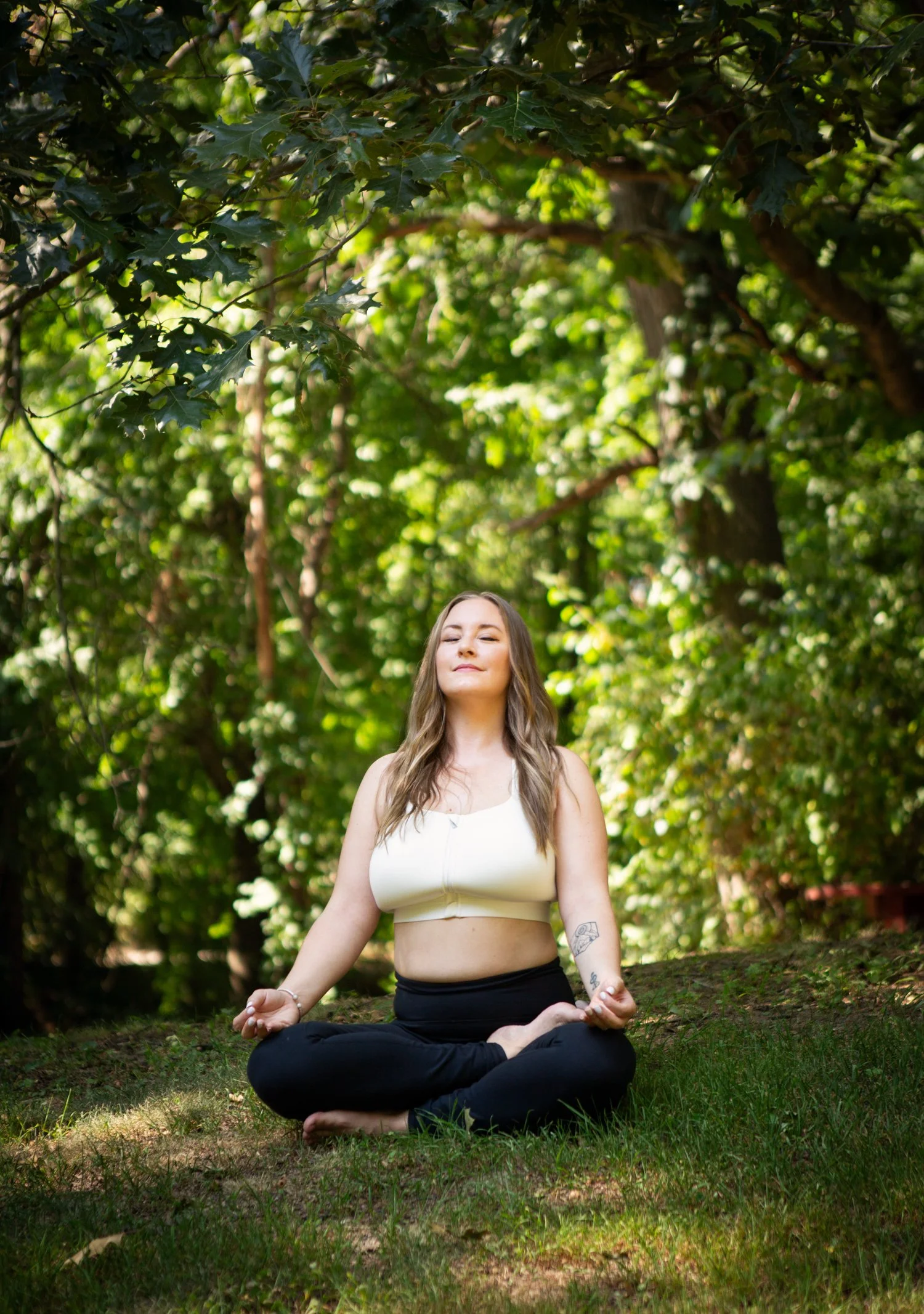 A woman practicing yoga outdoors in a forest, sitting cross-legged on the grass with a peaceful expression.