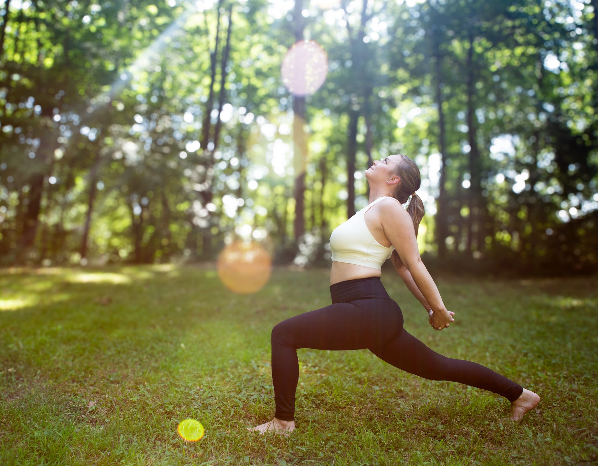 A pink yoga mat on the ground in a forest with green trees and foliage surrounding it, and a small water body visible in the background.