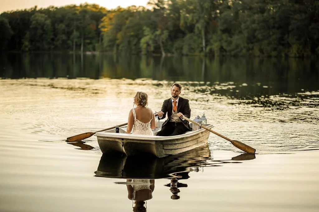 A couple dressed in wedding attire rowing a small boat on a calm lake during sunset, surrounded by trees with fall foliage.